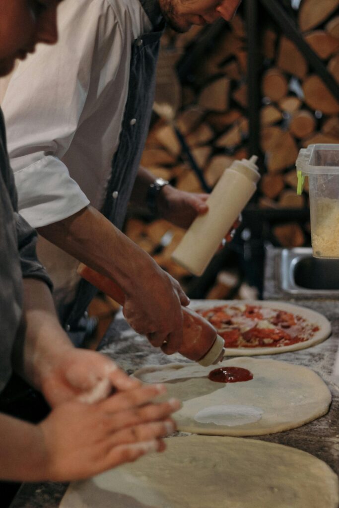 Two chefs skillfully prepare pizza, spreading dough and adding tomato sauce in a kitchen.