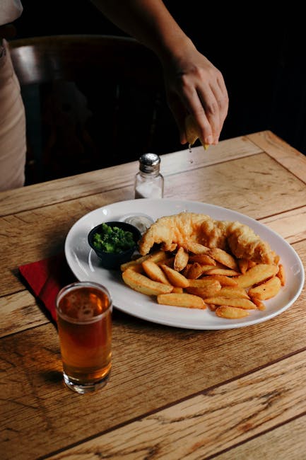 A plate of fish and chips with a beer on a wooden table, garnished with mushy peas and lemon.